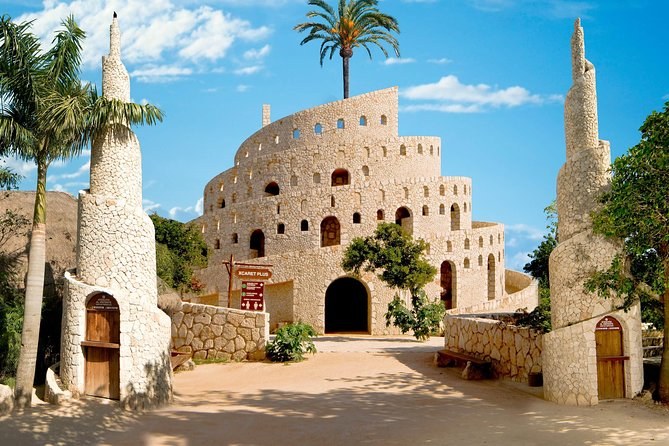 A pale stone building with a domed hall and two towers, palms in front of it